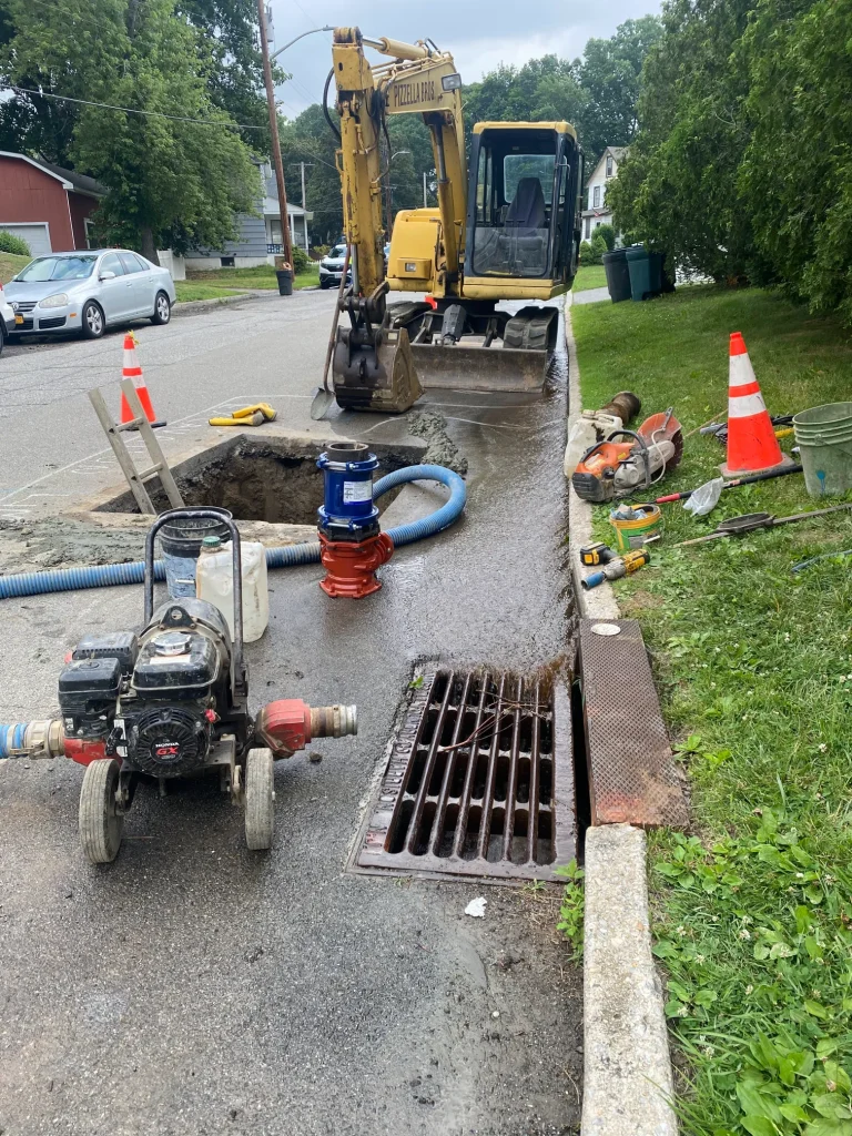 Backhoe next to open water main hole with a pump in front.